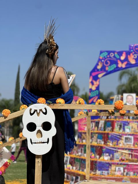 Catrina de espaldas con un tocado dorado de espigas y un rebozo azul, frente a un altar de retratos