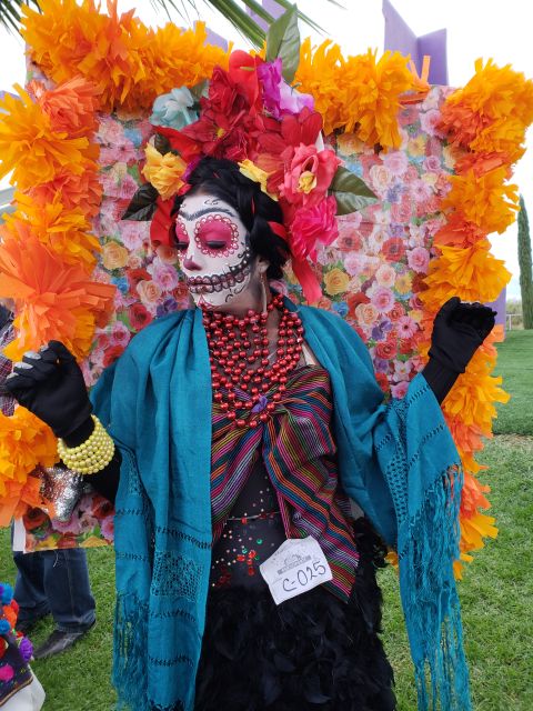Participante del concurso de disfraces caracterizada de catrina, con rebozo turquesa y collares rojos, frente a un marco de flores de papel
