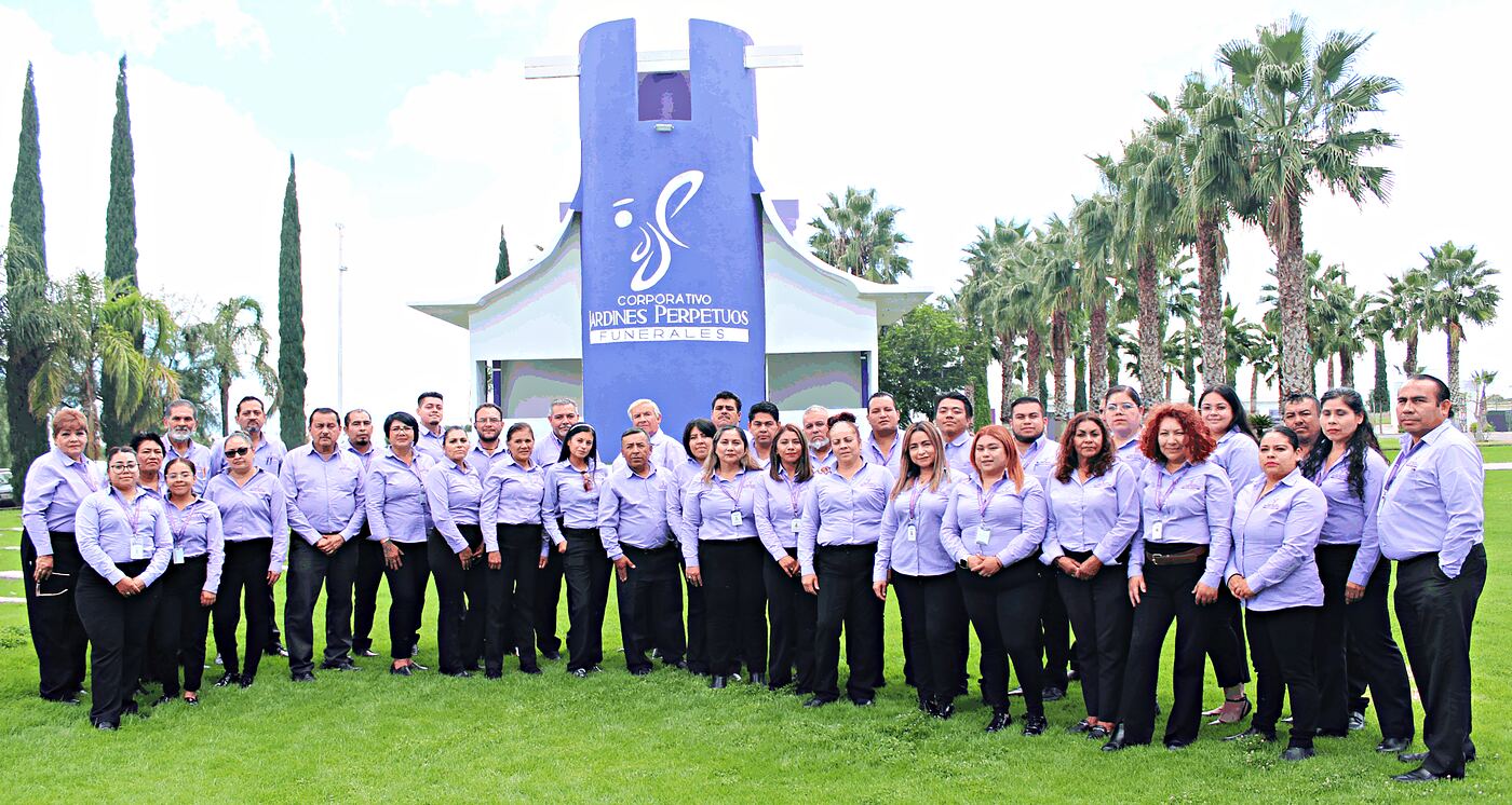 El equipo de Jardines Perpetuos, unas cincuenta personas con camisa lila, posando en el césped frente al acceso del cementerio